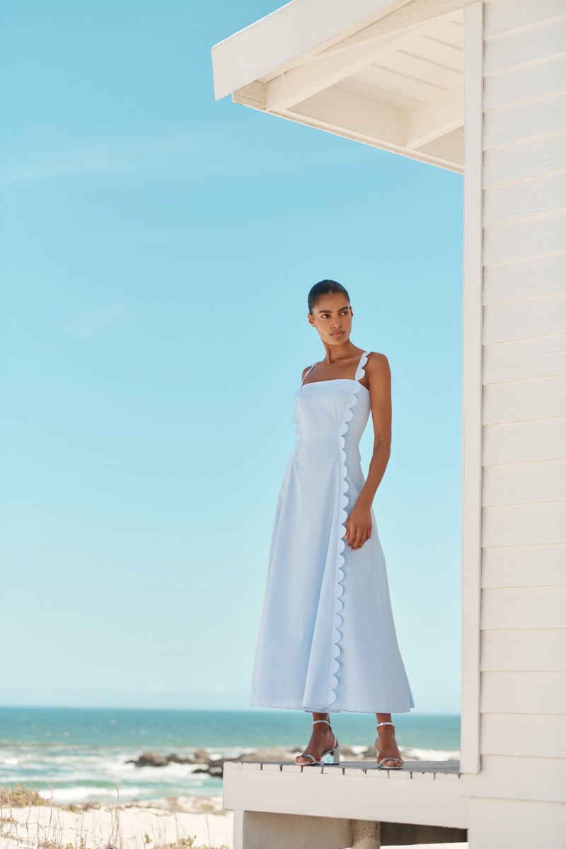 Wiggy Kit | Apron Dress (Blue Seersucker) | Model wearing maxi dress in light blue with beach in background