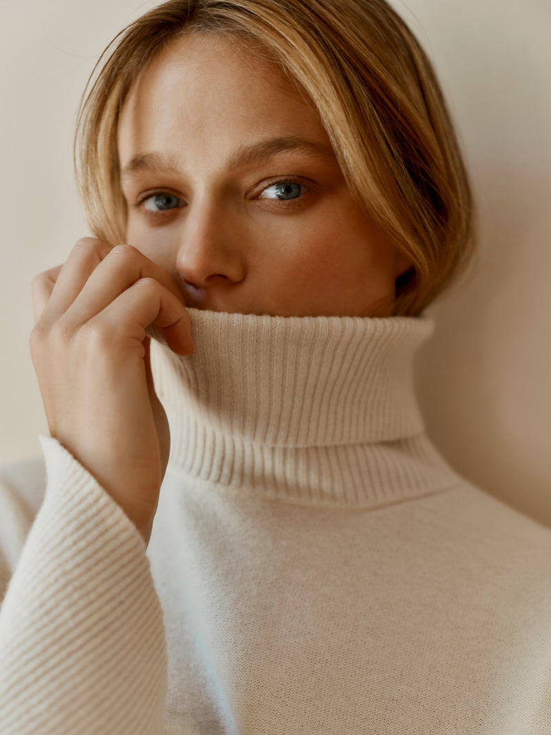 A model wearing the Wiggy Kit Inside Out Turtleneck in Ivory pulling the collar over their mouth.