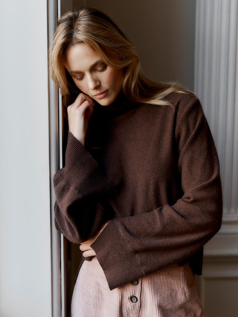 A model wearing the Wiggy Kit Inside Out Turtle Neck (Chocolate) looking down and leaning on a wall next to a window.