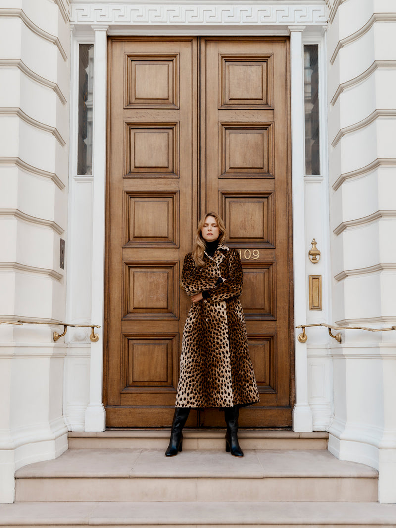 Wiggy Kit model wearing the Mercer Coat with both arms folder stood outside a grand front door.