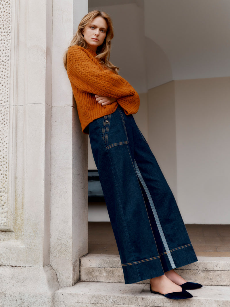 Wiggy Kit model wearing The Inside Stripe Jean and Slouch Fisherman's Crew in orange leaning in a grand doorway, arms crossed.