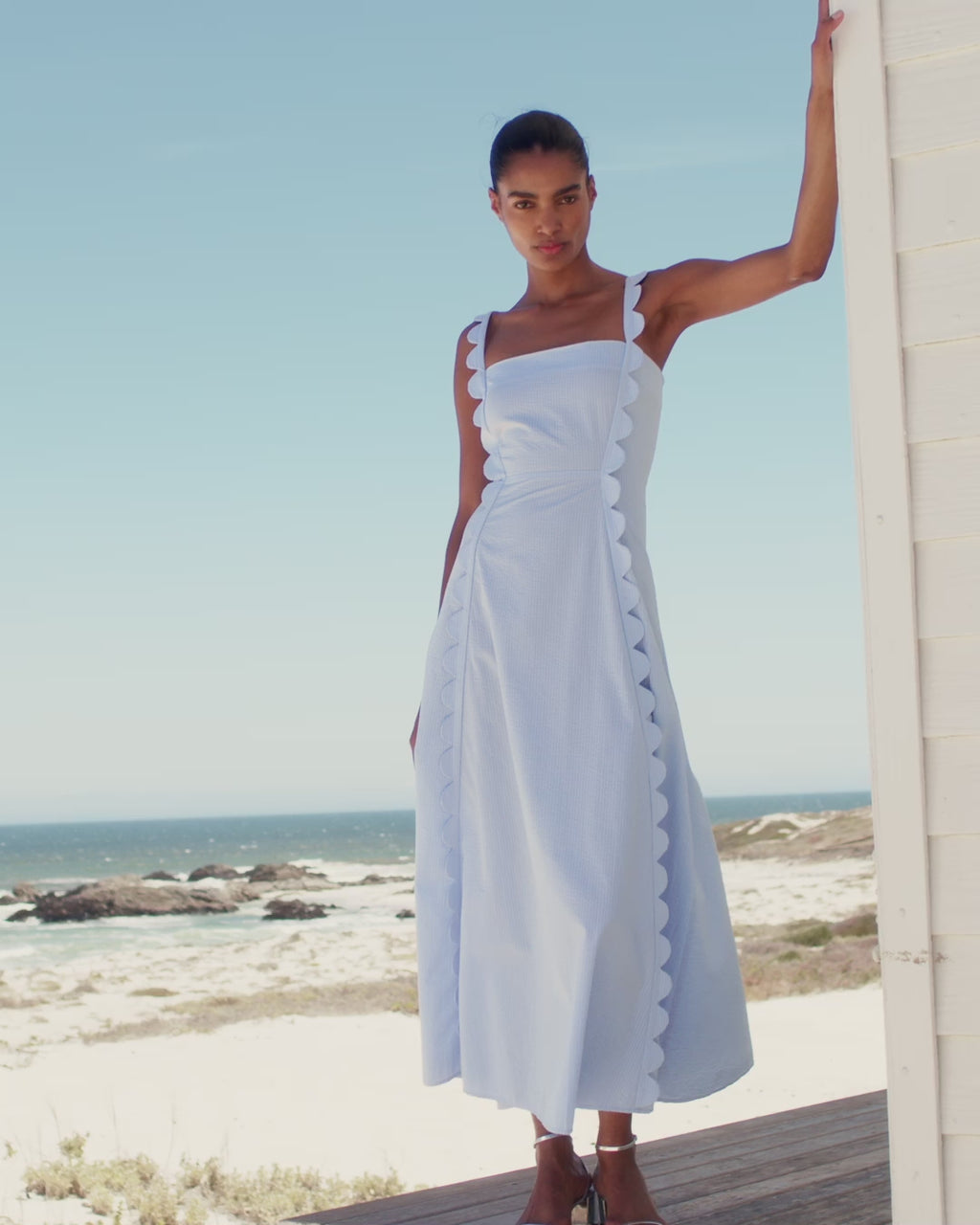 Wiggy Kit | Apron Dress (Blue Seersucker) | Model wearing maxi dress in light blue with beach in background